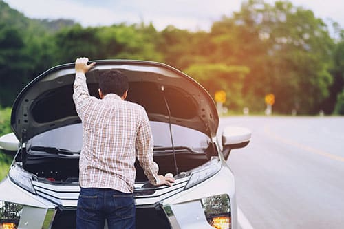 Man looking under roof of broken down car on the side of the road.