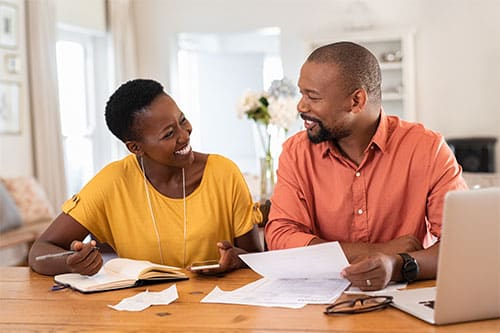 Man and woman sitting next eachother at surface in home discussing and smiling while laptop, documents and notebook rest in front of them.
