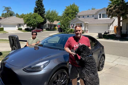 Couple and dog standing with new Tesla car sitting in driveway of neighborhood.