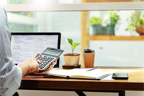 Person sitting at desk in office with laptop, coffee cup, plant and notebook in front of them while holding calculator in hand.