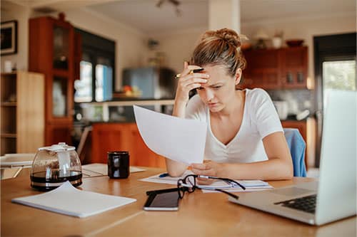 Woman with a frustrated lok on her face sitting at large surface surrounded by various items looking at paper in her hands.
