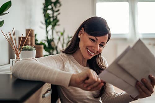 Happy woman resting on credenza while looking at sealed envelopes in her hand.