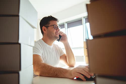 Man working at desk at computer surrounded by shipping boxes while holding phone up to his ear.