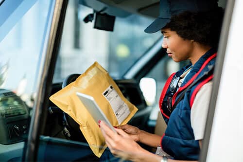Mailwoman sitting in the front of mailtruck looking at label on package and using phone in other hand.