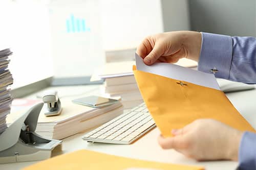 Person pulling document out of manila envelope at desk with keyboard, stacks of paper and office supplies surrounding them.