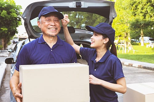 Elderly mailman and assistant standing with boxes on busy street with trunk door of car open.