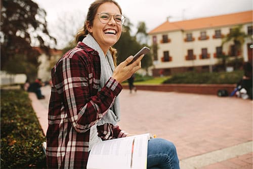 Happy woman sitting on ledge of flower bed holding phone in hand with text book open on her lap.