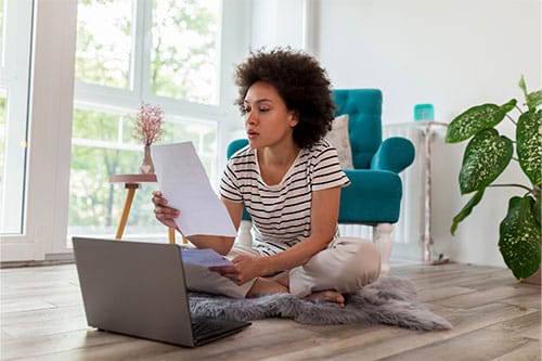Woman sitting on blanket on floor looking at documents in hands with laptop in front of her.