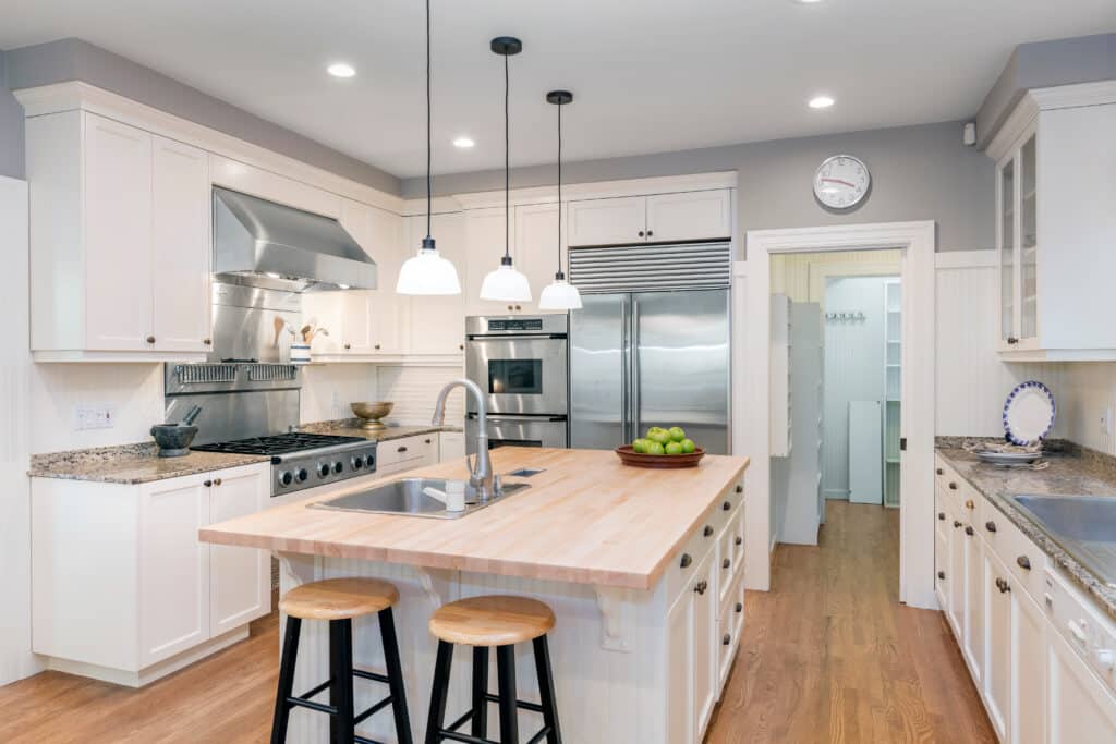 A modern kitchen with white cabinets, stainless steel appliances, and a butcher block island with two stools. Pendant lights hang above, and a bowl of apples sits on the counter, creating a welcoming and bright atmosphere.