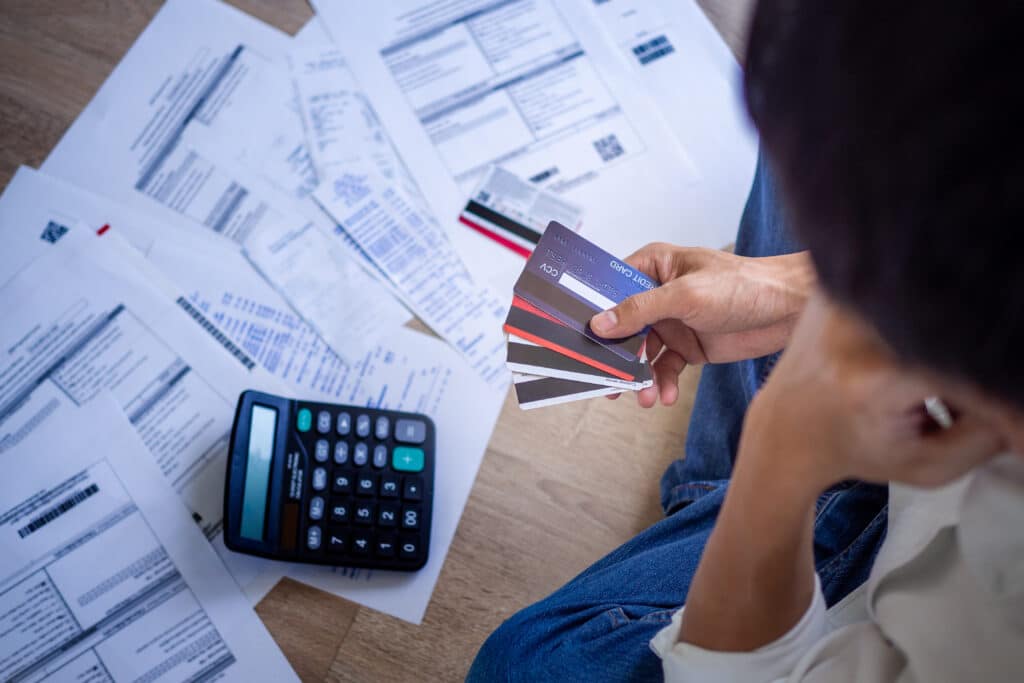 A person seated on the floor holds several credit cards, surrounded by scattered bills and a calculator, conveying financial stress and concern.