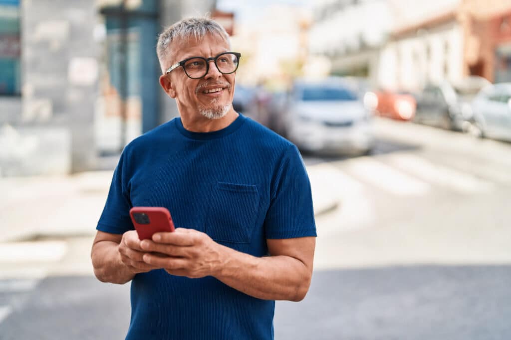 Man in glasses and blue shirt smiles while holding a red smartphone on a city street